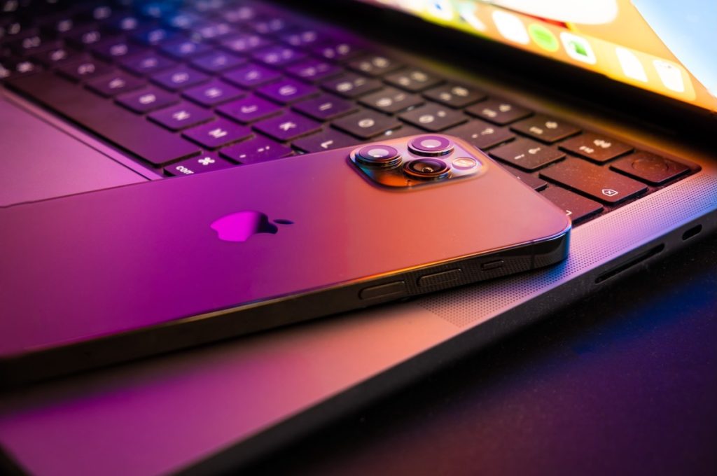 A suite of government hacking tools targeting iPhones is now being used by cybercriminals MacBook pro 2021 half open with Iphone 13 lit by retina display