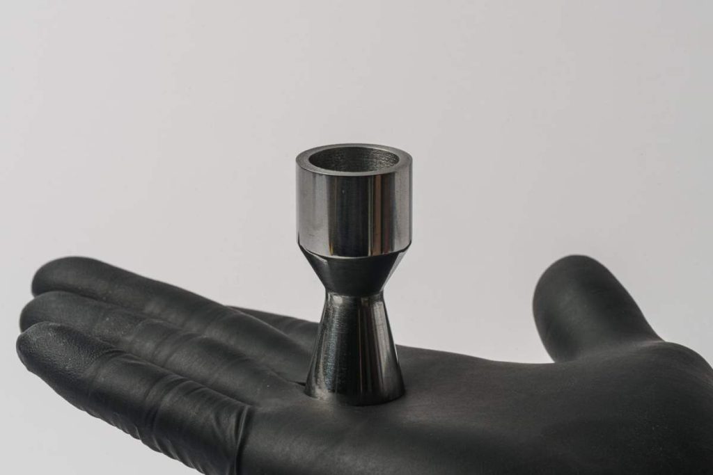 A gloved hand holds a metal part shaped like a rocket nozzle.